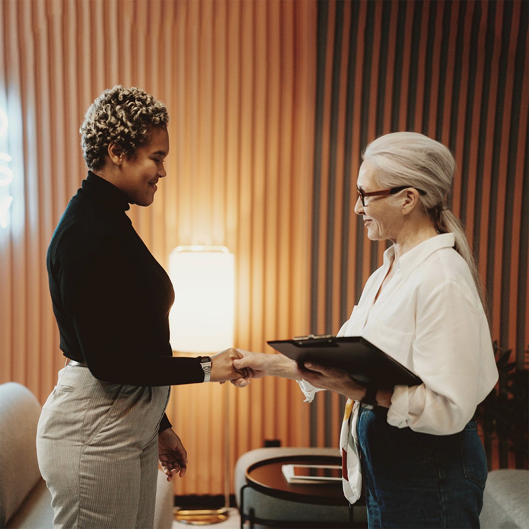 A black woman with curly hair wearing a black turtleneck and a white woman with long white hair wearing a white business blouse shake hands in an office