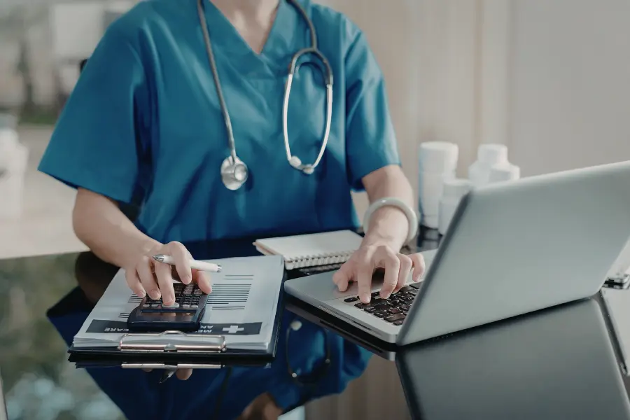 Healthcare professional in blue scrubs using AI hospital tool on a laptop while reviewing patient records on a clipboard.