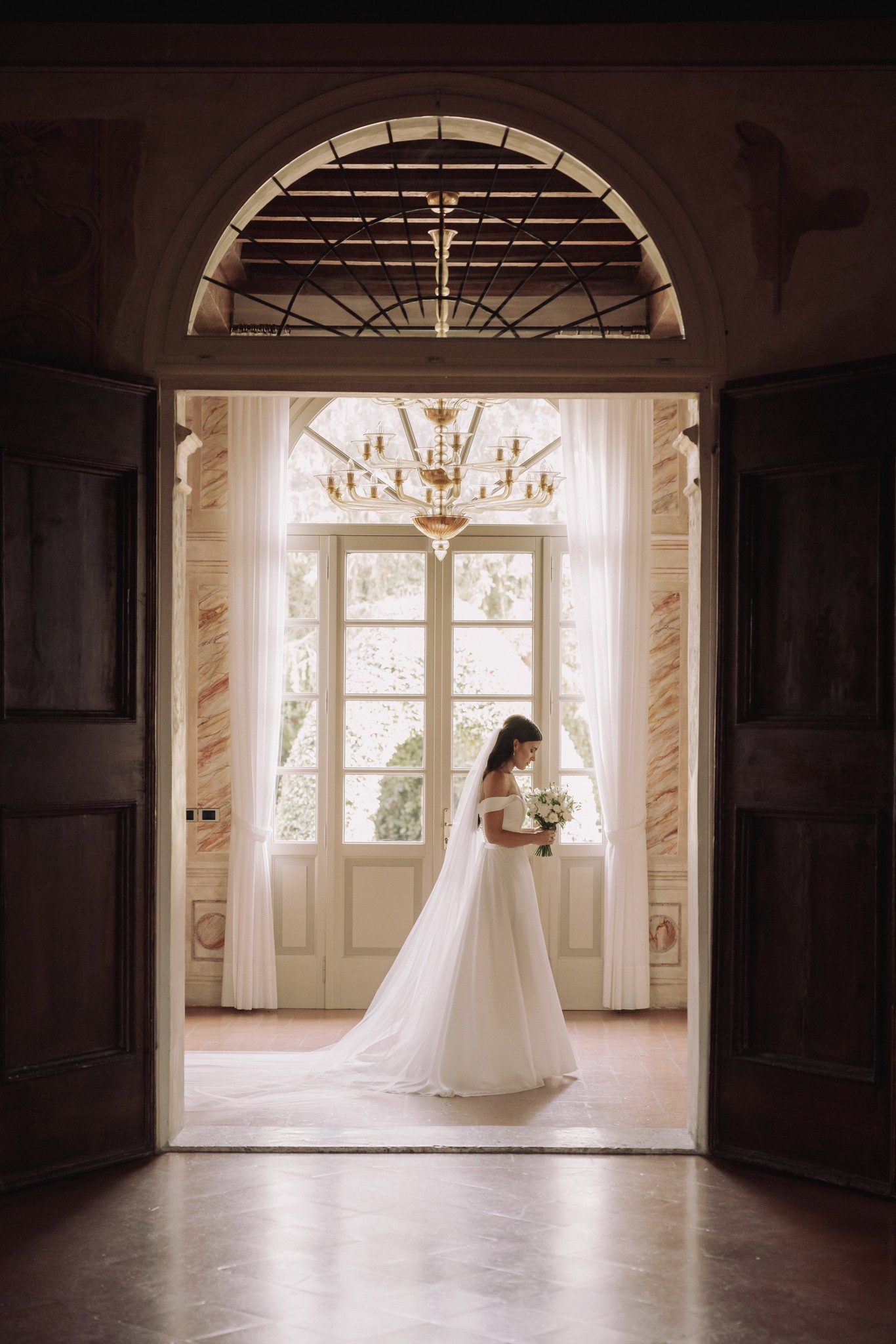 Sposa con bouquet bianco tra le porte aperte di Villa Negri, velo lungo, luce naturale nel corridoio del matrimonio