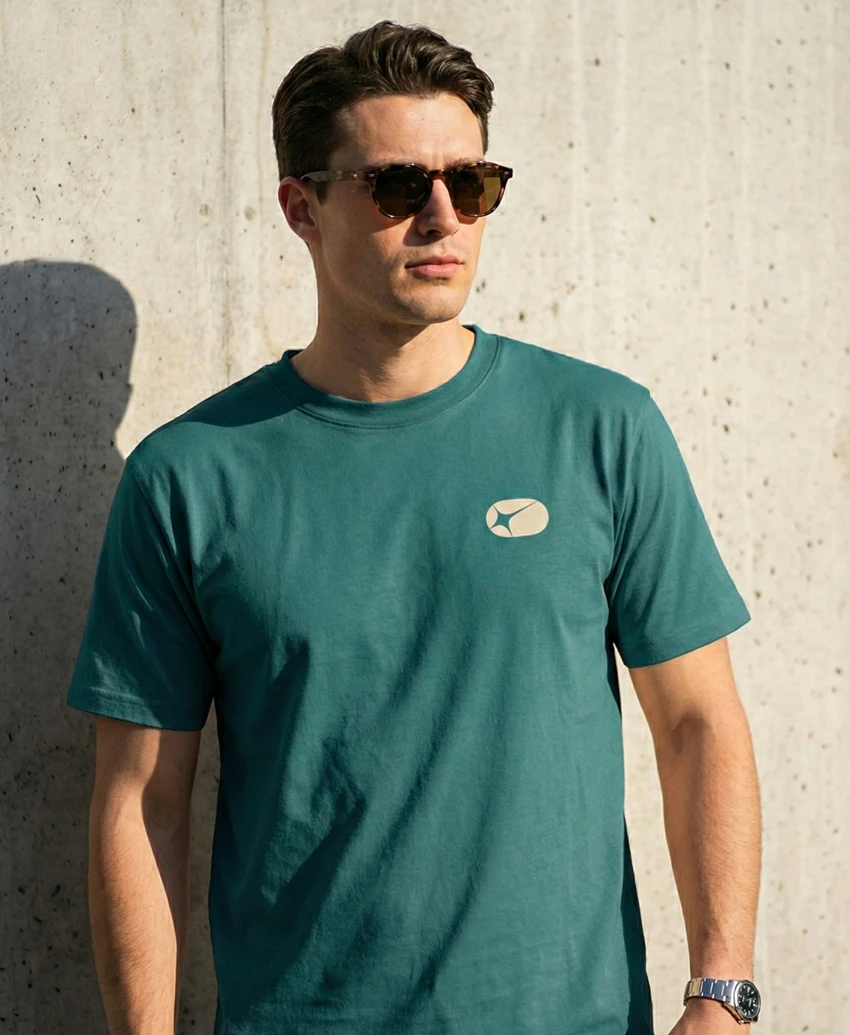 A man in sunglasses wearing a forest teal T-shirt with a small sand Action Replay icon on the chest.