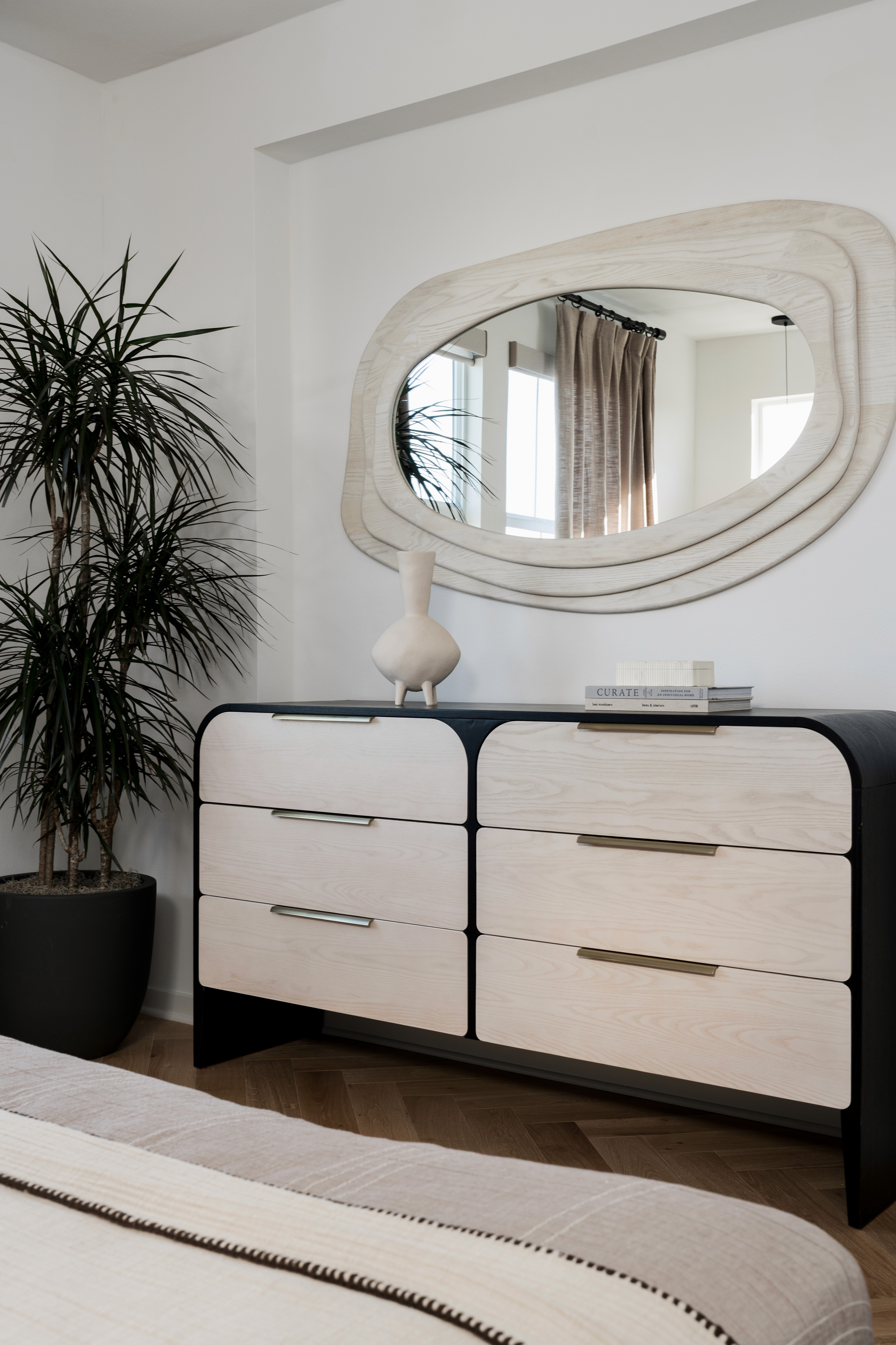 Rounded, black framed dresser with natural wood colored drawers, and brass finishes with large potted plant and modern wall mirror above dresser