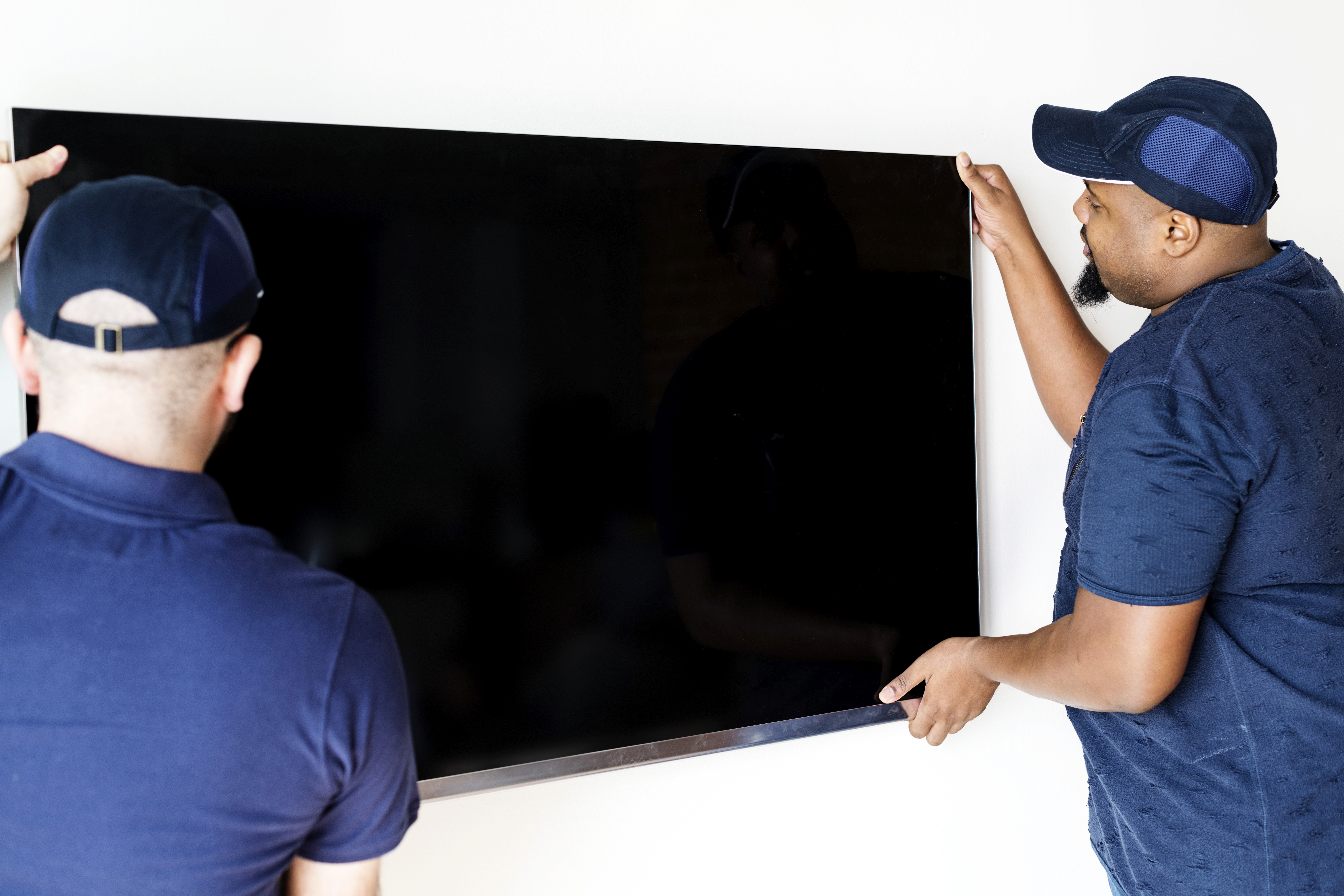Two men in blue shirts and caps are carefully unmounting a large flat-screen TV on a white wall. The scene conveys focus and teamwork.