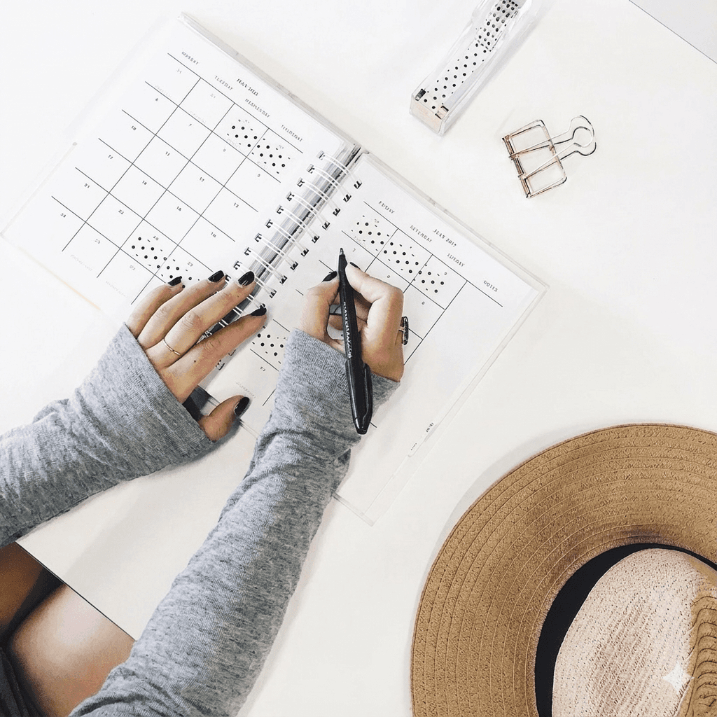 Top-down view of a woman with long grey sleeves and black nail polish writing in a spiral-bound planner on a white desk, surrounded by a straw hat, a gold paper clip, and a ruler.