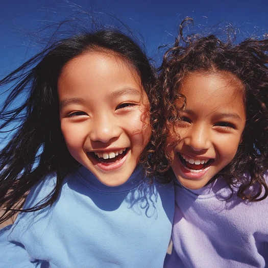 Two young children smiling brightly, wearing light blue and lavender shirts, against a clear blue sky. Their hair is windswept, evoking joy and playfulness.