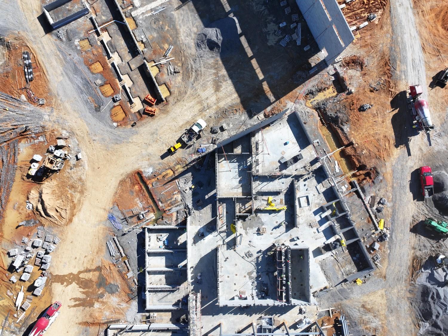 Drone overhead shot of active consturction site with partially completed walls of a commercial building with no roof showing the base concrete floor with construction equipment and vehicles on site.