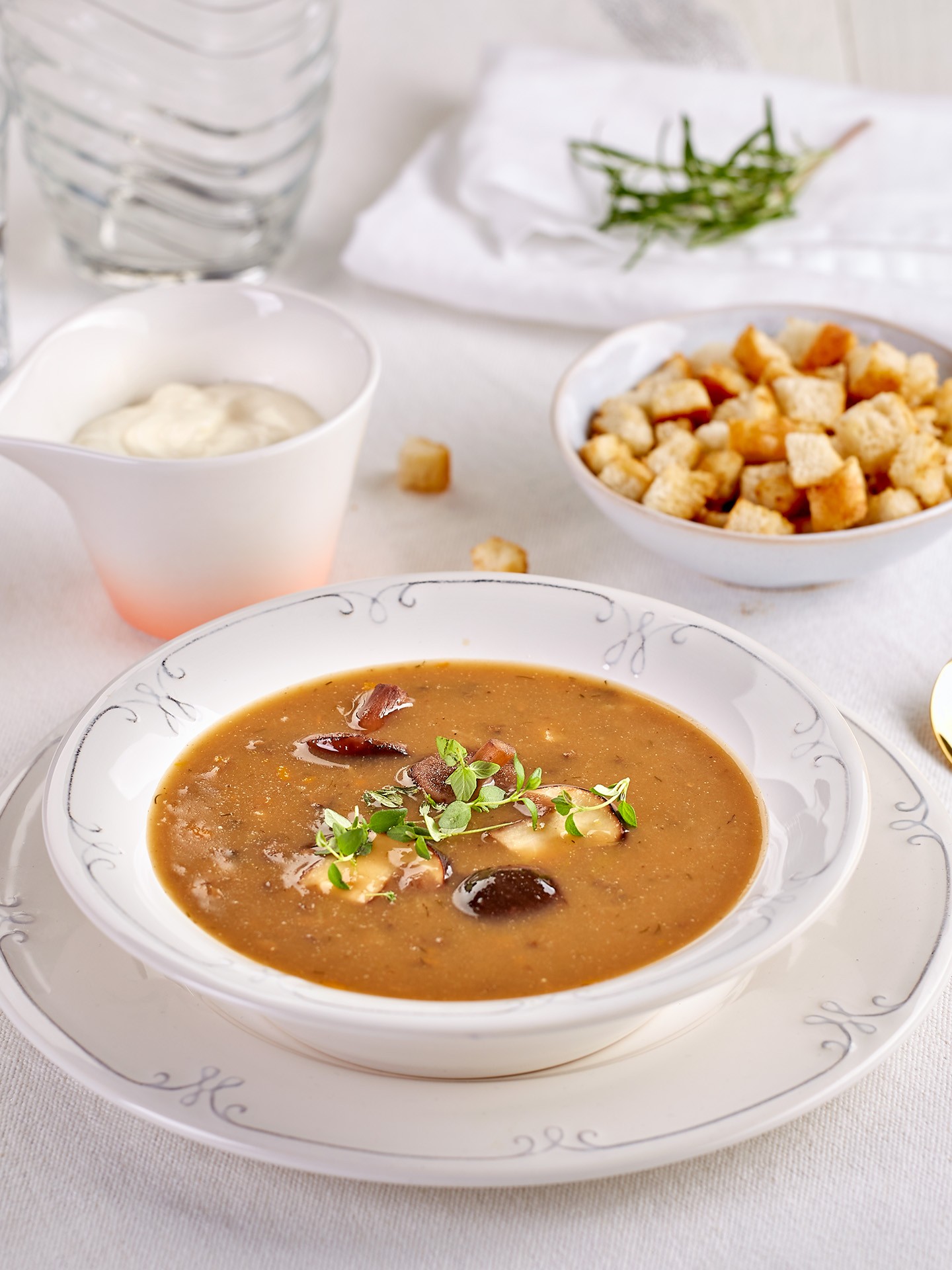 A creamy mushroom soup garnished with fresh herbs and croutons is served in an elegant white bowl, accompanied by a small dish of croutons and a cup of creme, all set on a white tablecloth with a sprig of rosemary and a glass of water in the background.
