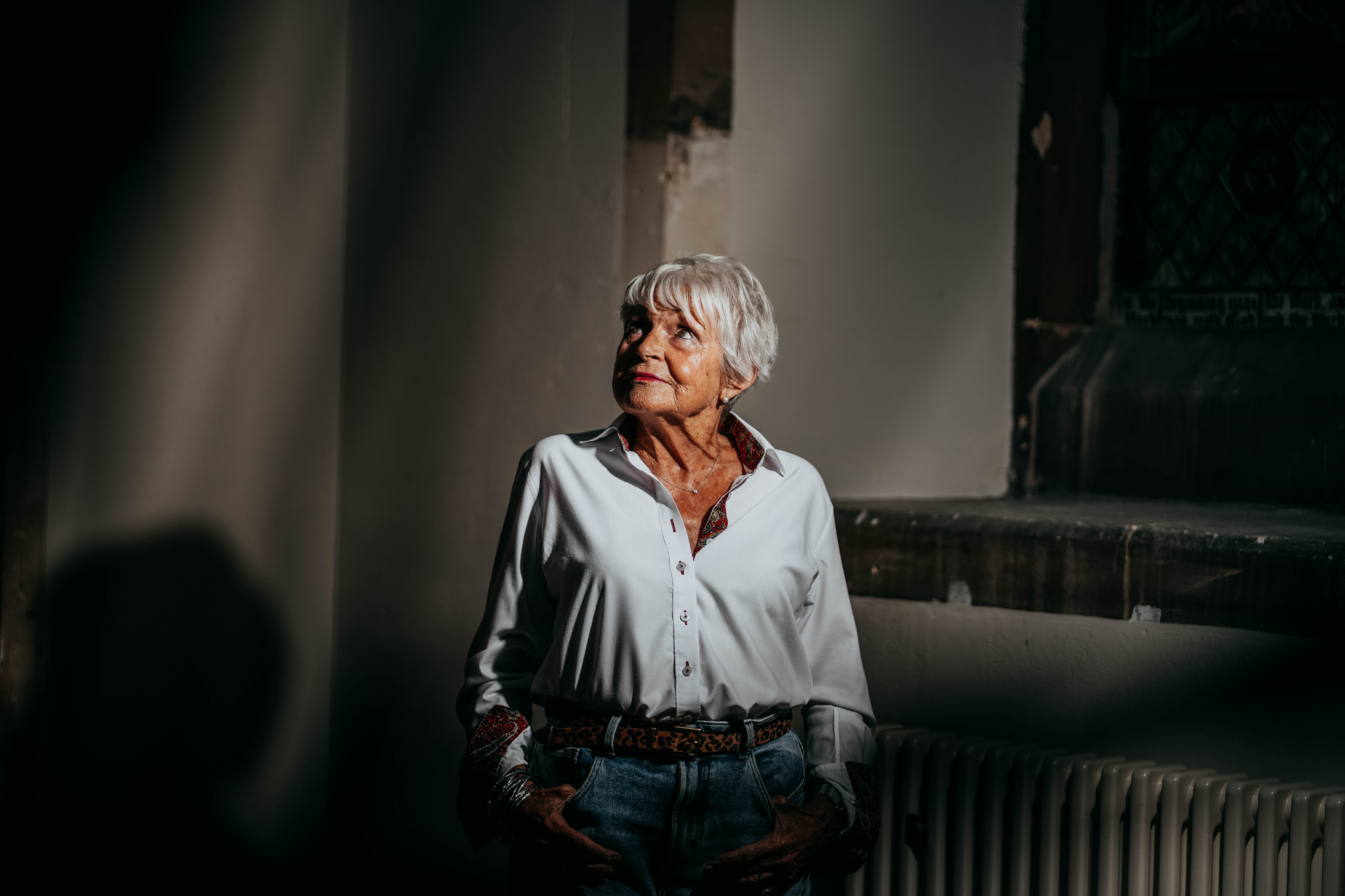 An older woman with short white hair, wearing a white shirt and jeans, stands in a beam of light, looking up and to the side with a contemplative expression inside a dimly lit building.
