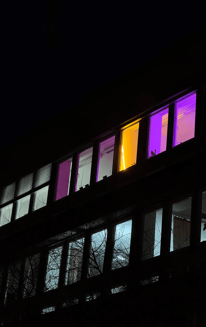 A nighttime exterior view of the new studio office in Zurich, featuring a row of windows glowing with vibrant purple and bright amber light. This visual serves as a "beacon" for the studio's new beginning, highlighting its presence within the urban creative landscape.