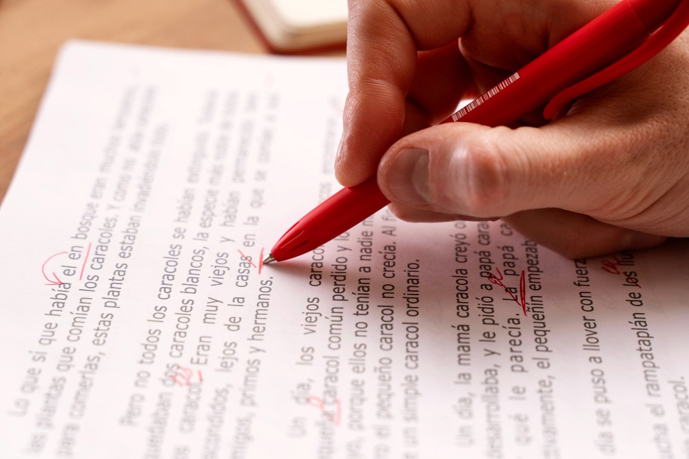 A hand holding a red pen is writing on a sheet of lined paper filled with text.