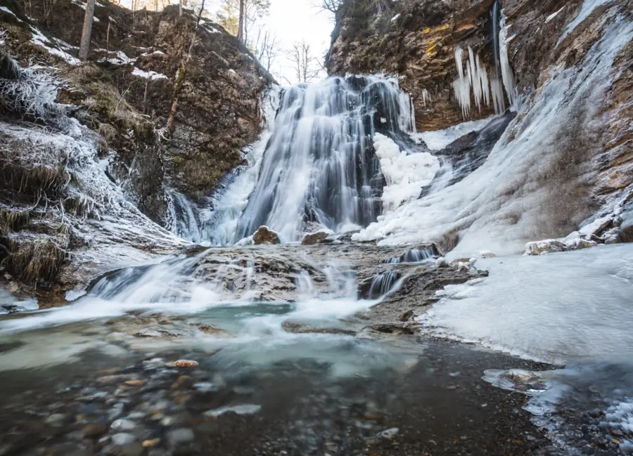  The multi-tiered Stegovnik Waterfall in Slovenia during winter, featuring rushing water cascading over rocky ledges surrounded by thick white ice formations and long icicles in a frost-covered forest canyon.