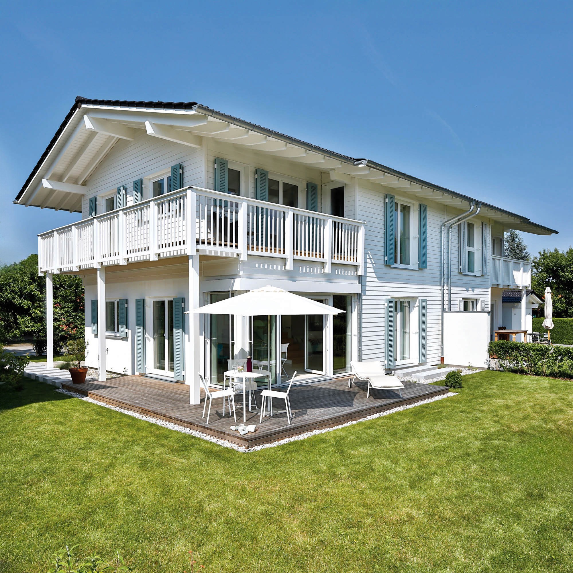 Weißes Holzhaus mit blauen Fensterläden, Balkon, Terrasse und Gartenmöbeln an einem sonnigen Tag.
