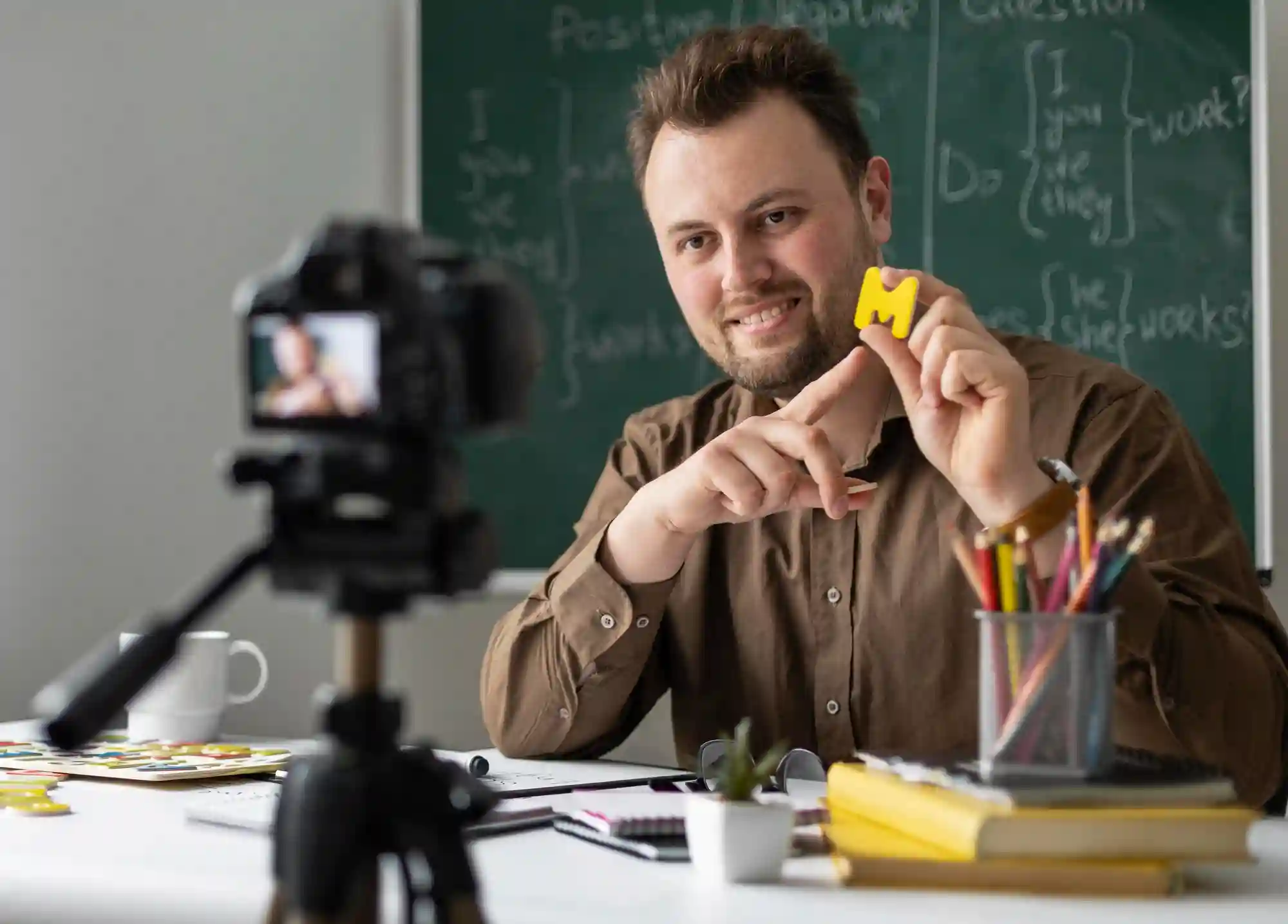 Male teacher records an educational video tutorial, holding a small object and sitting in front of a chalkboard.