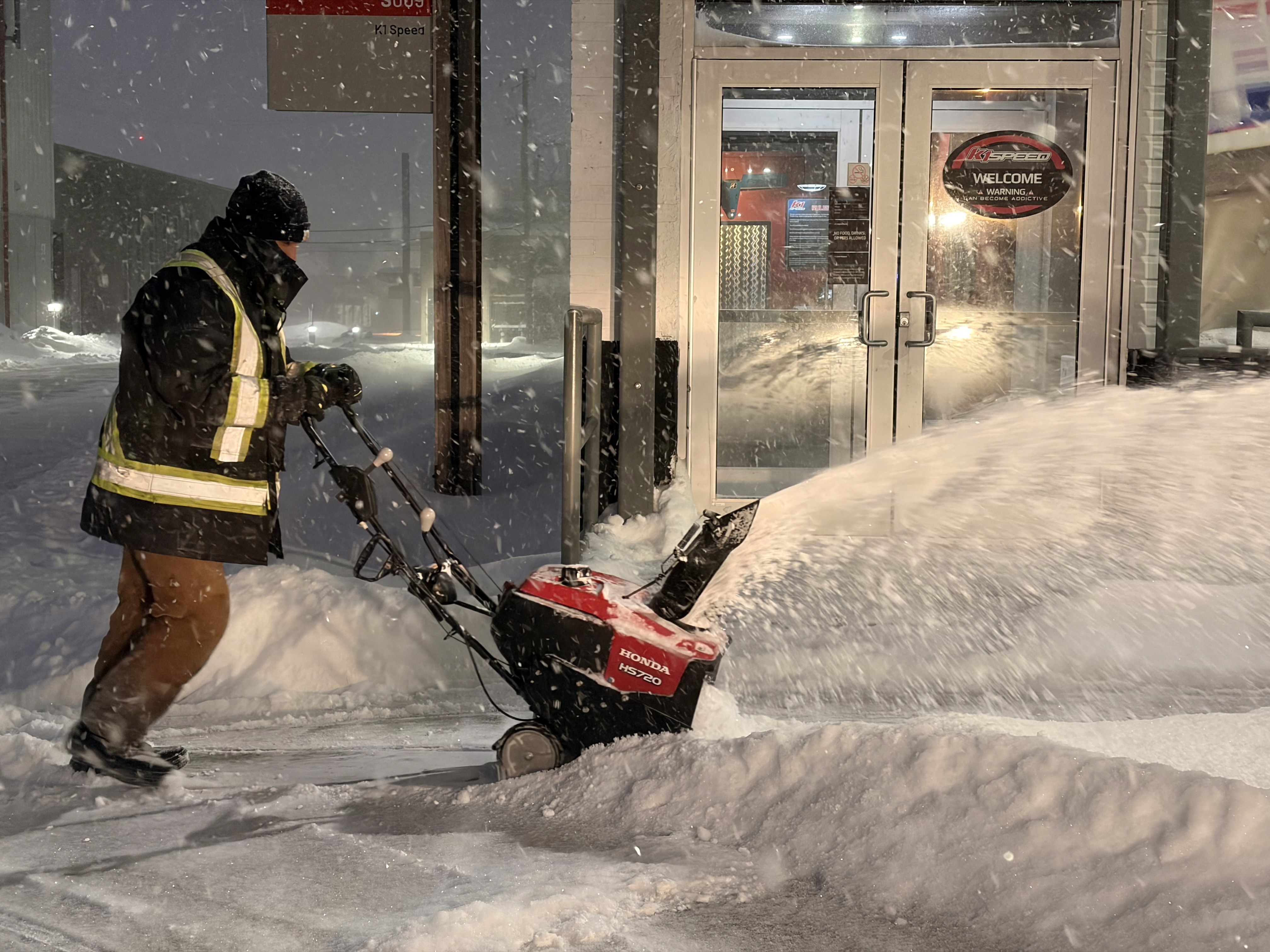 A person in a reflective jacket shovels snow in a city street, with piles of snow and a building in the background.