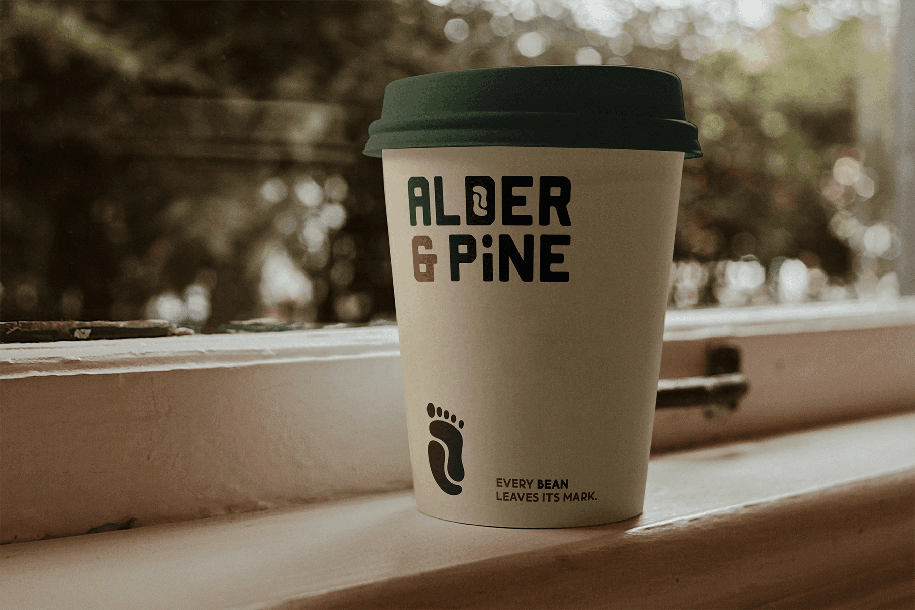 Graphic design mockup of a coffee cup for Alder & Pine Coffee Roasters — modern, clean wordmark style logo of a footprint and a coffee bean with a bold decorative sans serif typography, designed by Will Lefkovich.