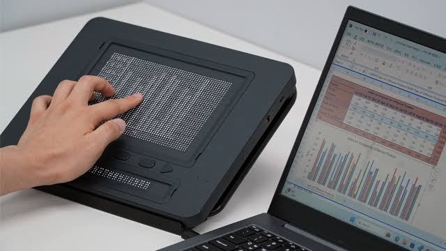 A person reads the raised-dot surface of Dot Pad beside a laptop displaying a spreadsheet and a bar chart.