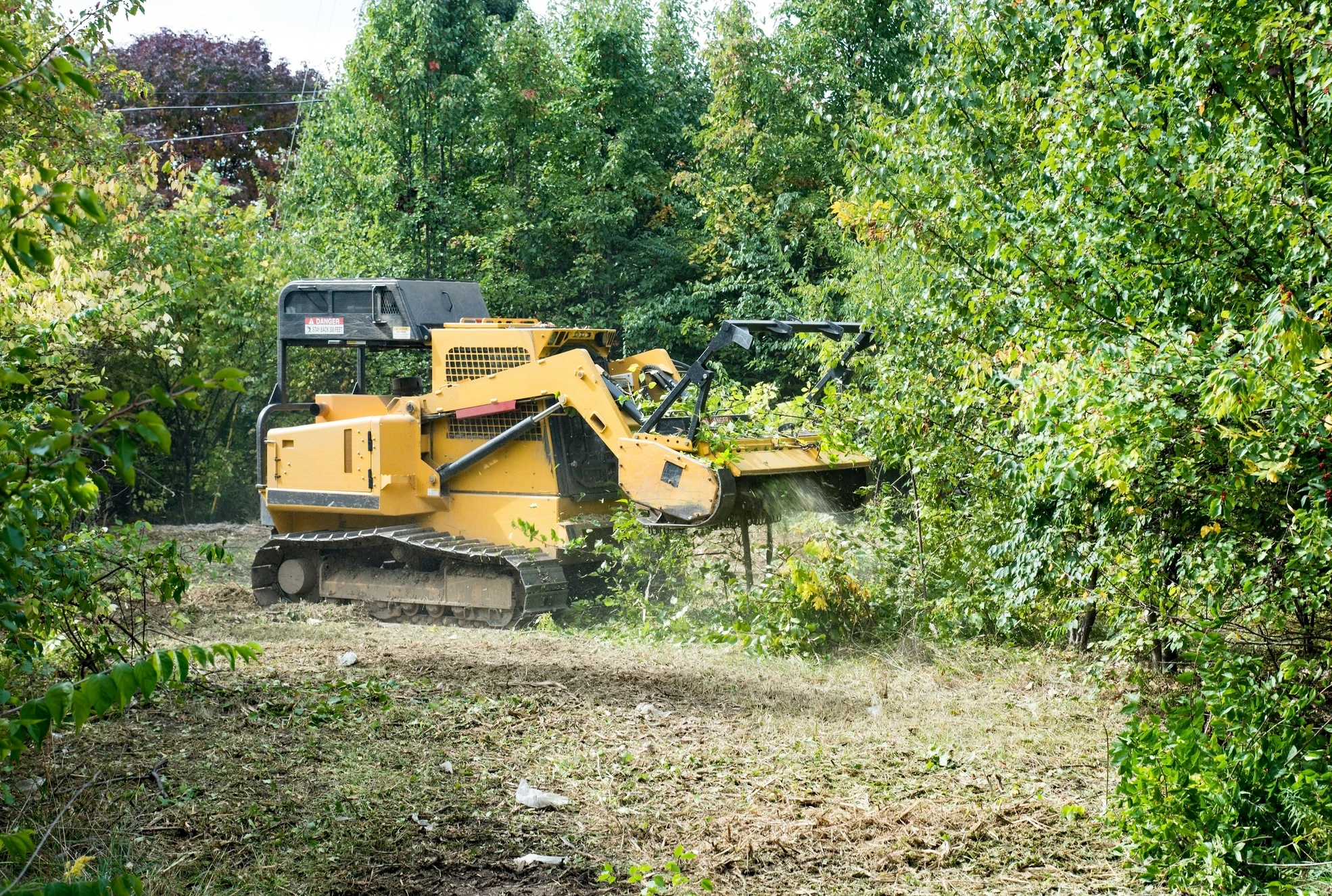 A yellow tracked vehicle with a rotating drum mulcher attachment is clearing brush and small trees in a wooded area. Debris is flying up as the machine works.