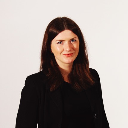 A woman with long brown hair and light skin is wearing a black blazer over a dark top. She is facing forward and looking directly at the camera with a slight smile. The background is a plain off-white or light beige color.
