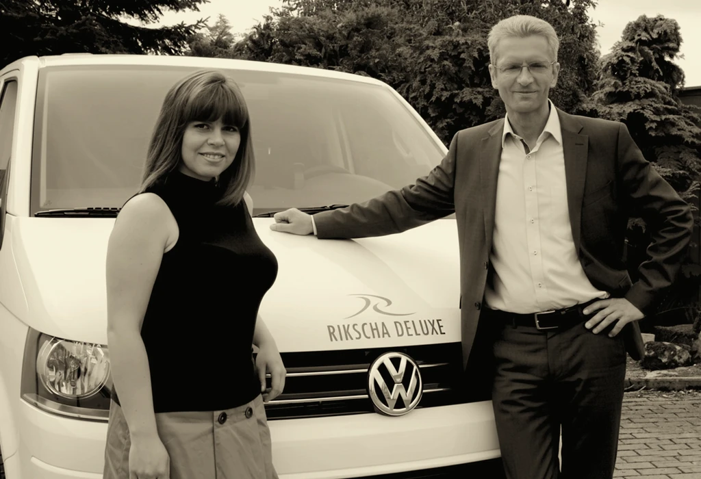 Ein lächelndes Paar steht neben einem weißen VW-Bus. Auf der Motorhaube des Busses steht „RIKSCHA DELUXE“.
