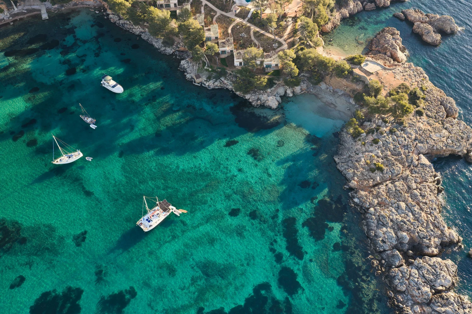 Eine Luftaufnahme einer türkisfarbenen Bucht mit mehreren Segelbooten und Yachten, die vor einer bewaldeten Steilküste ankern.