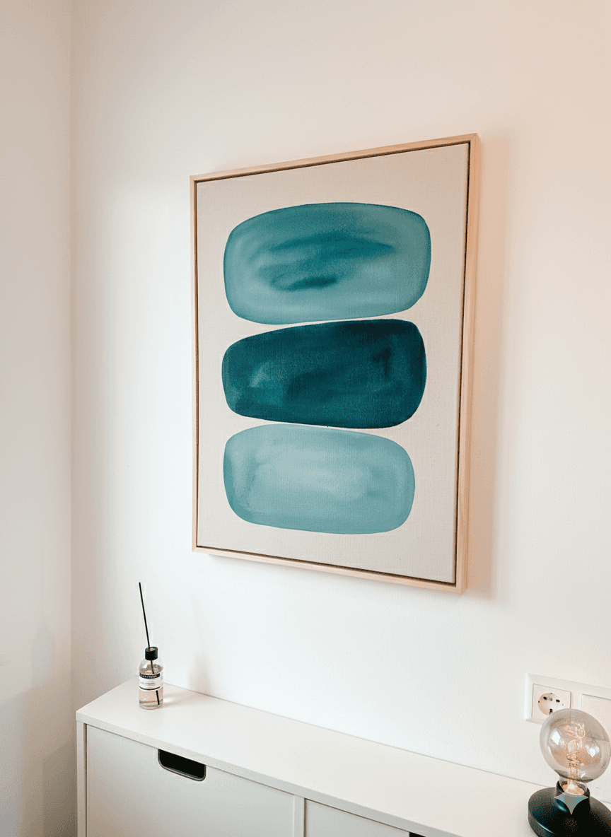 A framed artwork with two large teal oval shapes hangs on a light wall above a white surface.
