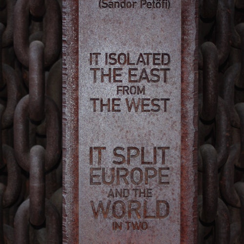 A vertical metal plaque with the text: "IT ISOLATED THE EAST FROM THE WEST. IT SPLIT EUROPE AND THE WORLD IN TWO." Chains are visible in the background.