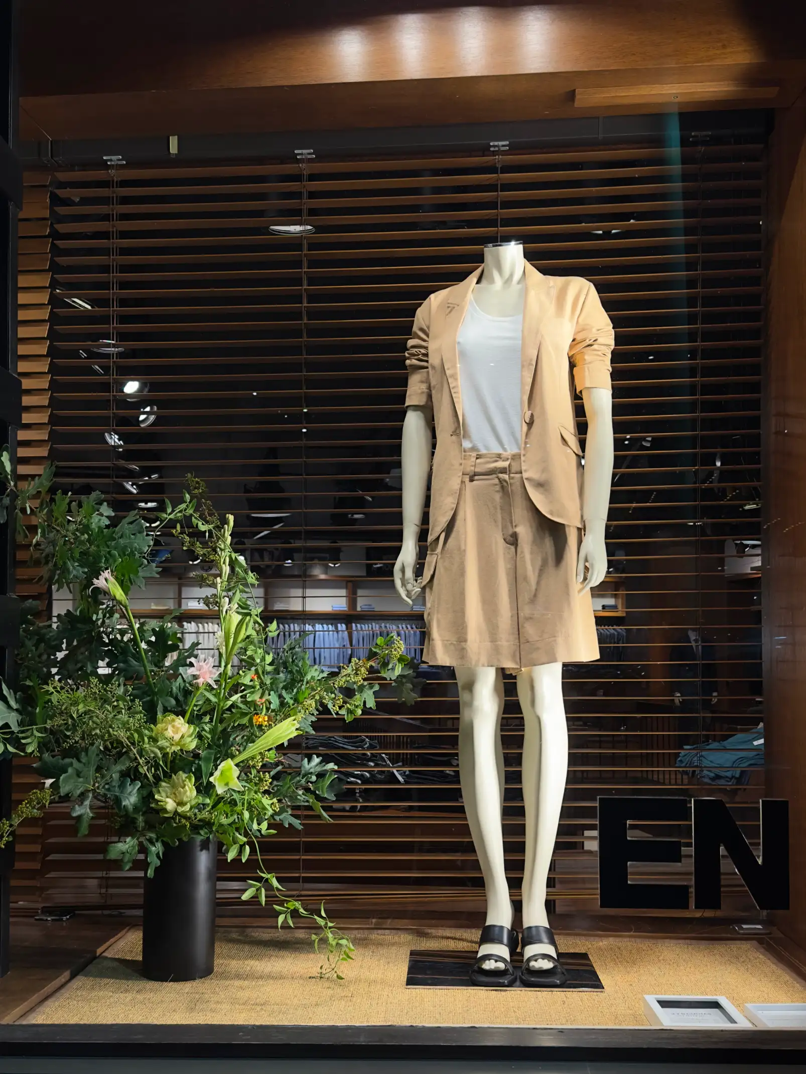 Vitrina de tienda con maniquí y traje beige junto a arreglo floral verde para diseño de vidriera sofisticada.