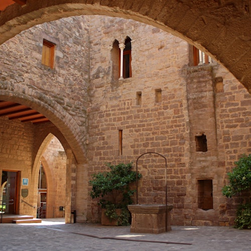 Stone courtyard with archways, a well, and greenery. Brick walls feature small windows and arched openings.