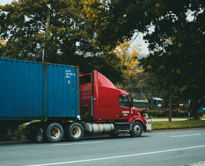 Red semi-truck hauling a blue shipping container parked on a roadside with trees in the background.