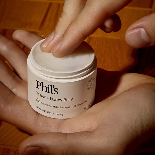 A lifestyle photograph showing a 50ml white VIVOMER tub of Phil's Tallow and Honey Balm held in a hand. The image highlights the ergonomic and retail-ready nature of this home-compostable skincare packaging.