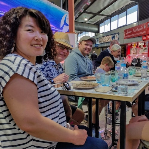 Group of people sitting around a table with drinks and plates inside a bustling indoor market or food court.