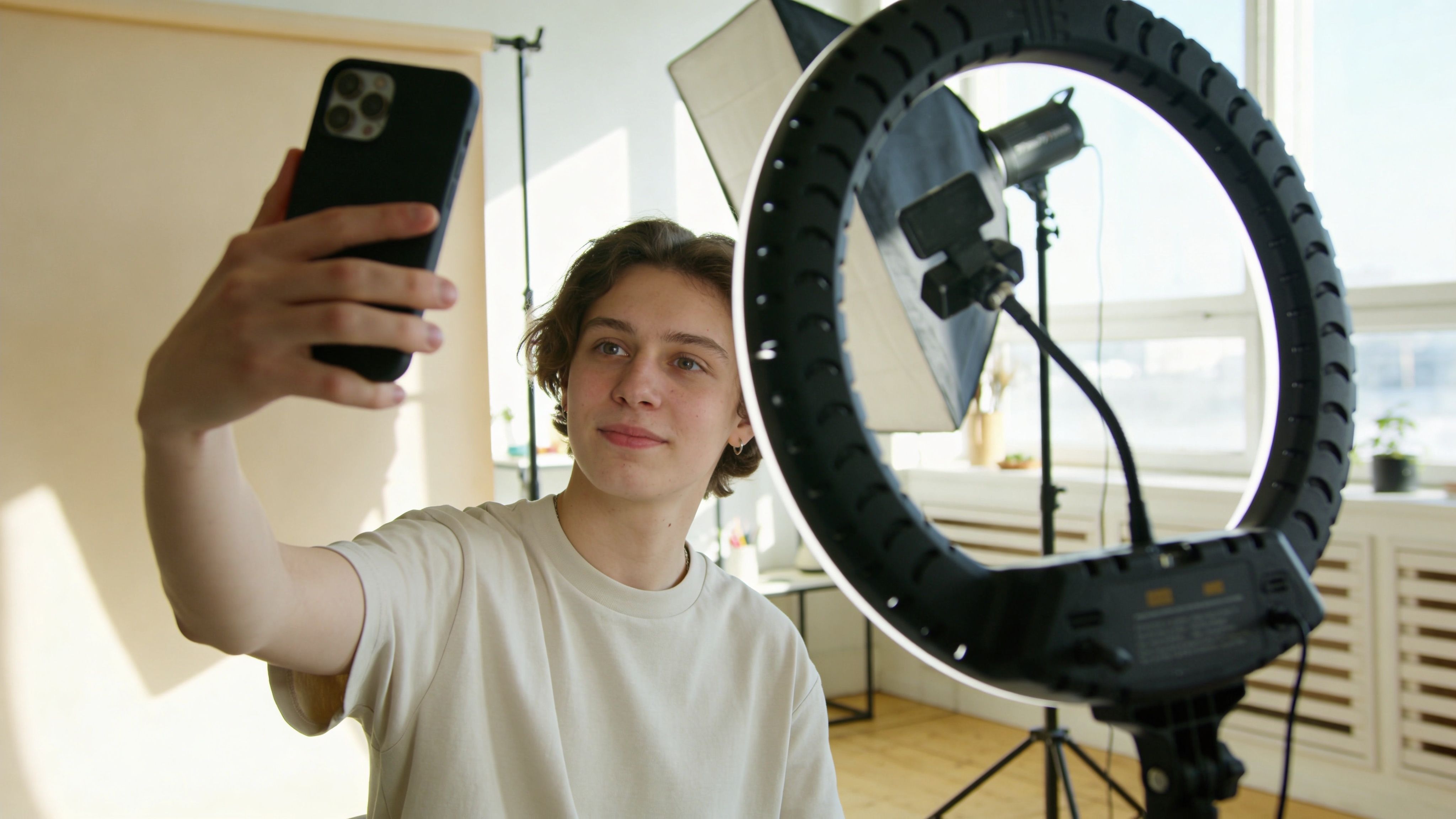 Ein junger Mann mit braunen Haaren nimmt in einem hellen Fotostudio ein Selfie mit seinem Smartphone auf.