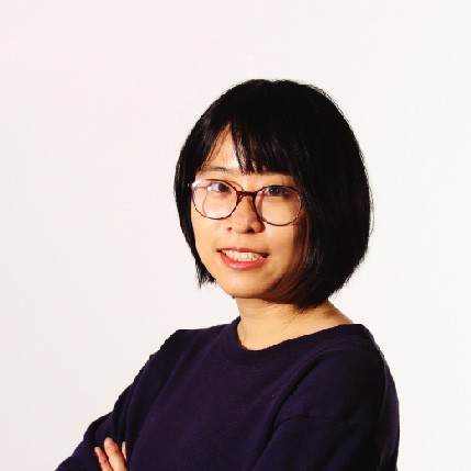 A person with short, dark hair and bangs is wearing a dark blue, round-framed top. Their arms are crossed over their chest. They are looking directly at the camera with a slight smile. The background is a plain white.