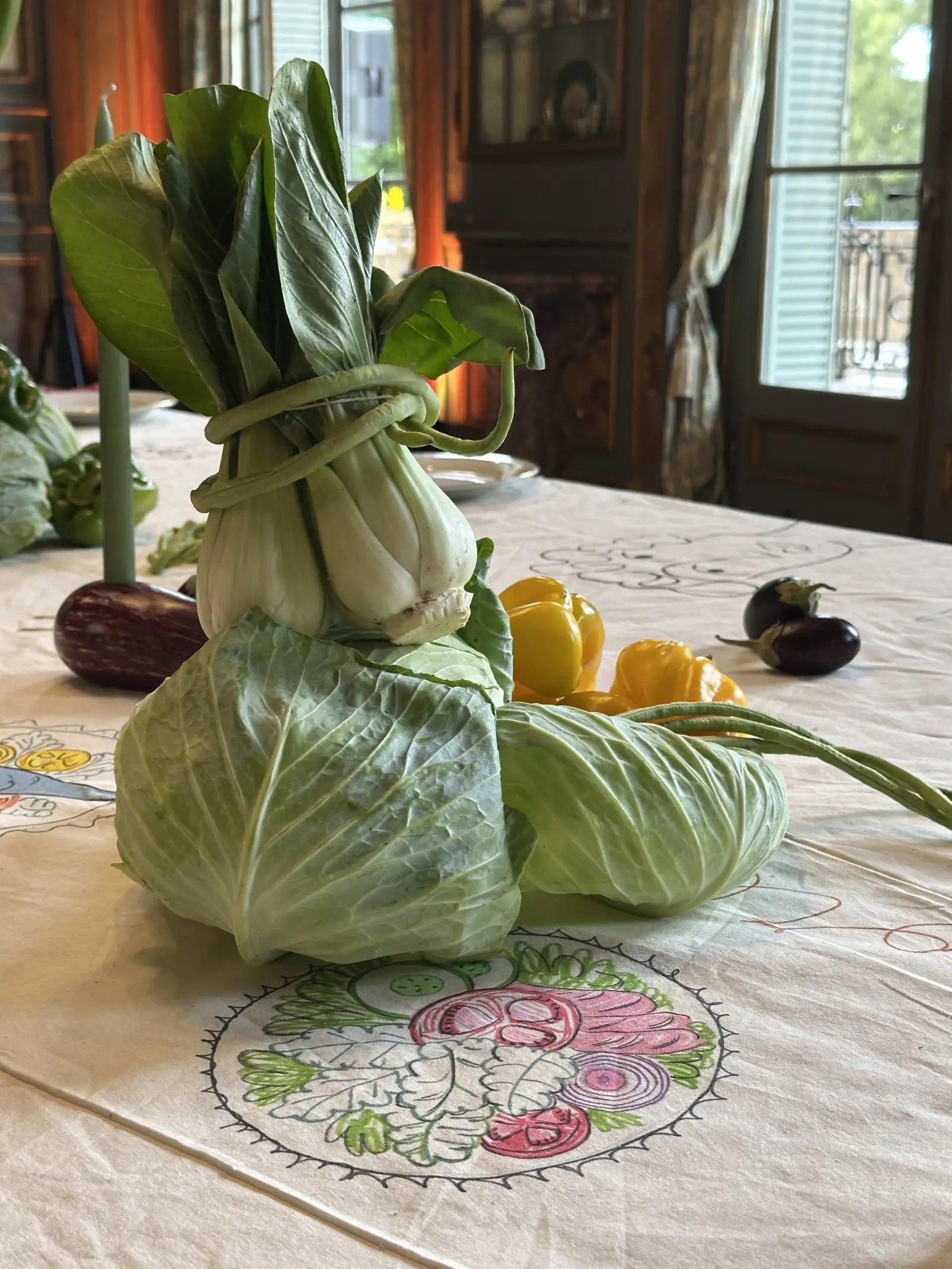 Composición decorativa de vegetales sobre un mantel ilustrado, con un repollo y un pak choi atados formando una figura, acompañados de pimientos amarillos y berenjenas en una mesa elegante junto a una ventana