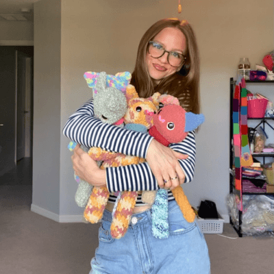 A person with a light beard, wearing an orange sweater, is smiling with a thoughtful gesture against a warm yellow background.