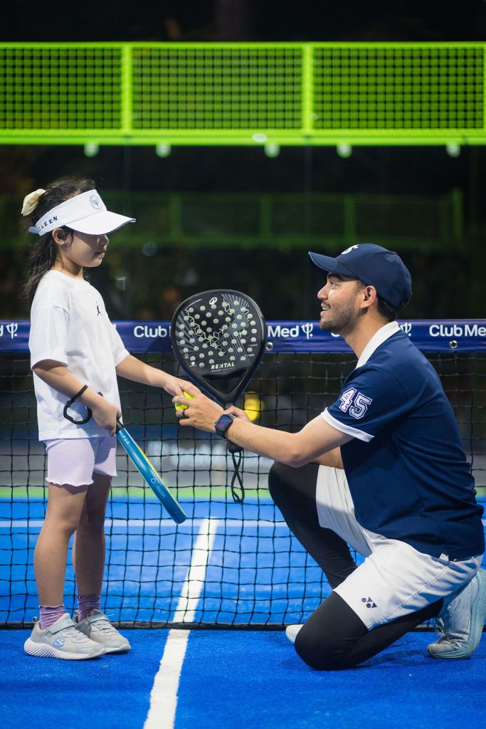 A young girl wearing a white cap and t-shirt stands with a paddle in hand, facing a smiling man in a blue sports outfit who is kneeling and holding a tennis ball. They are on a brightly colored tennis court with a net in the background.