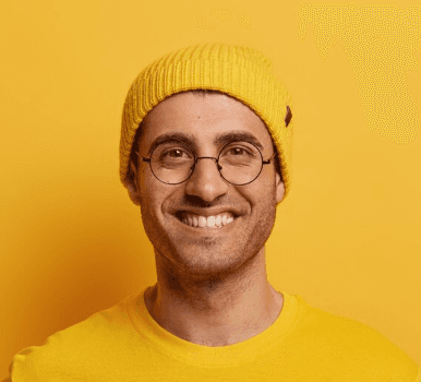 A person wearing round glasses, a yellow beanie, and a yellow t-shirt stands smiling against a vibrant yellow background.