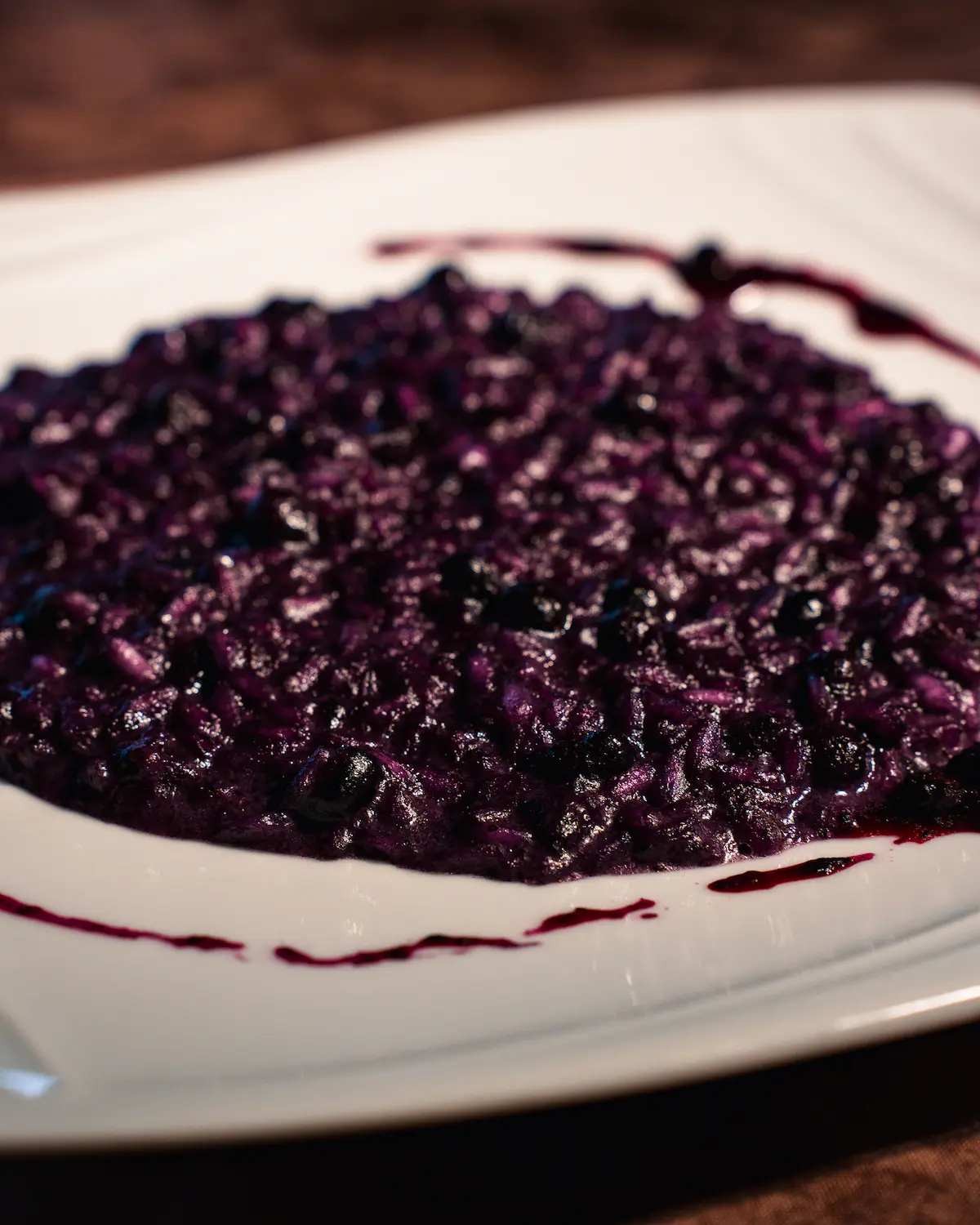 A group of people stand under olive trees, engaged in conversation, in a sunlit orchard with a distant farmhouse visible, illustrating a serene countryside gathering.