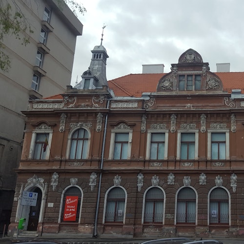 装飾的な石細工とアーチ型の窓、赤い瓦屋根が特徴の華麗な茶色の建物。隣接するのは灰色の多層建物です。