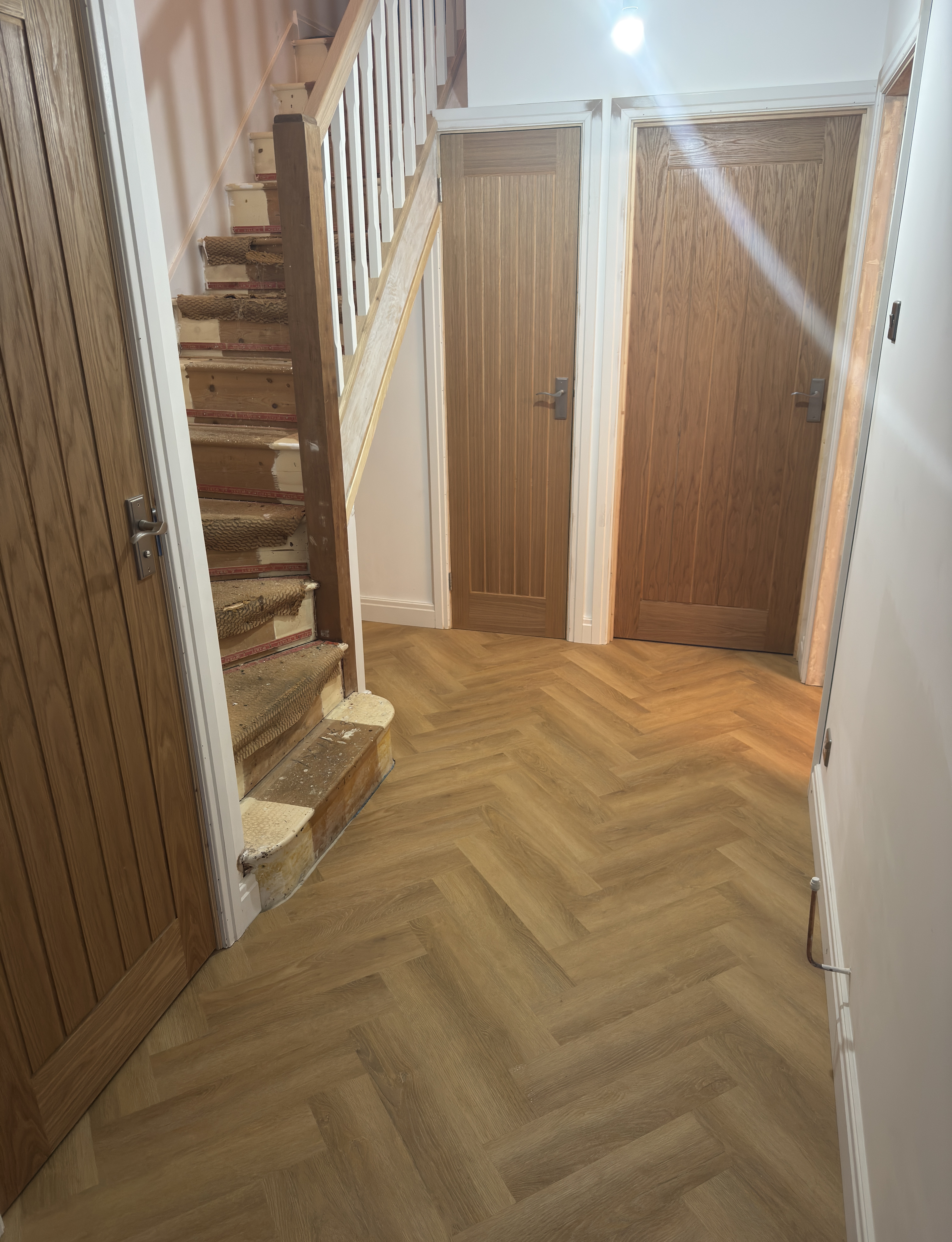 A modern hallway features vibrant teal and orange geometric and leaf patterns, with glossy tiled floors, a large rectangular light fixture, and a small shelf holding a potted plant, all leading to a large, frosted window at the end.