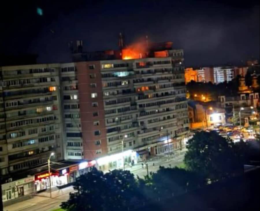 Apartment building at night with flames and smoke visible on the rooftop, viewed from across a city street.