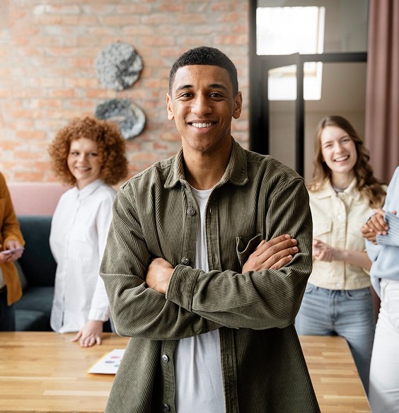 A confident man stands with arms crossed, smiling, surrounded by diverse colleagues in a bright office setting.