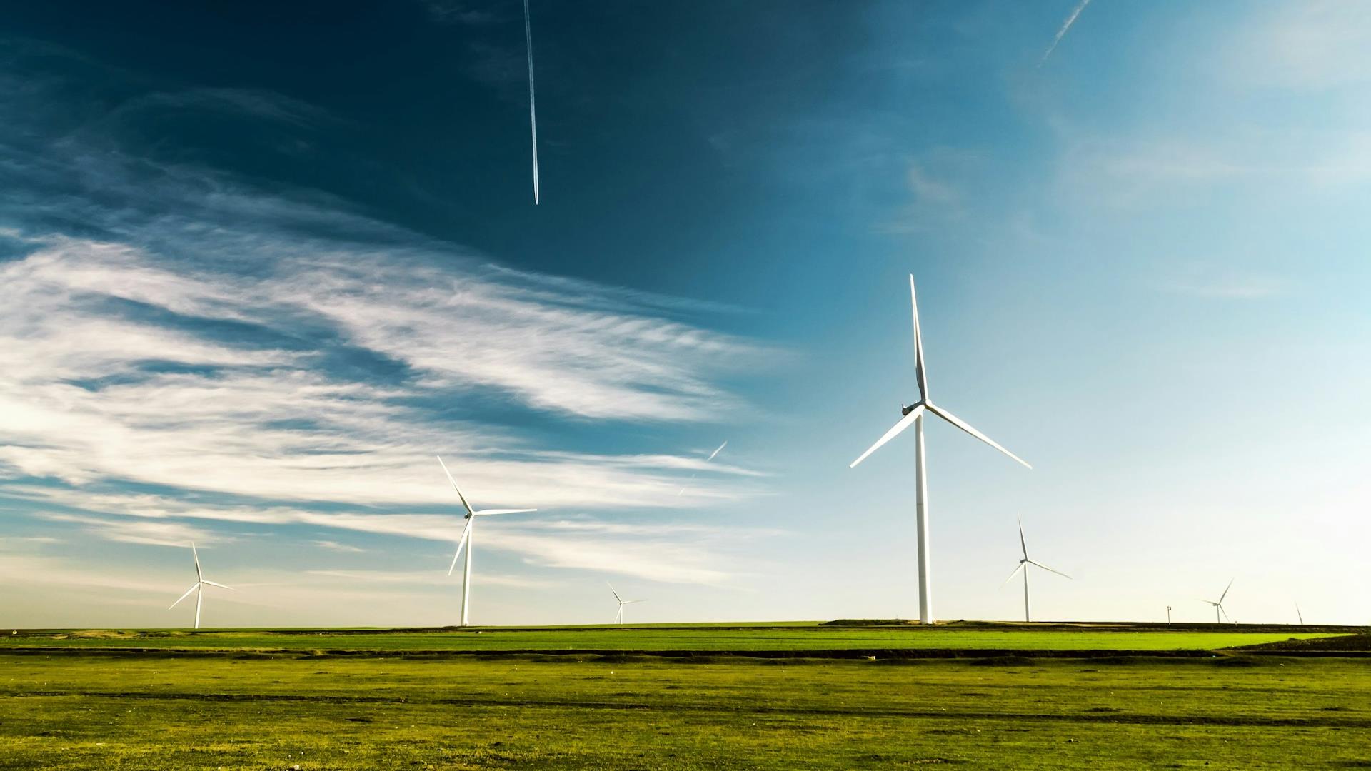 Wind turbines stand tall on a lush green field under a blue sky with wispy clouds and contrails, conveying a sense of tranquility and sustainability.