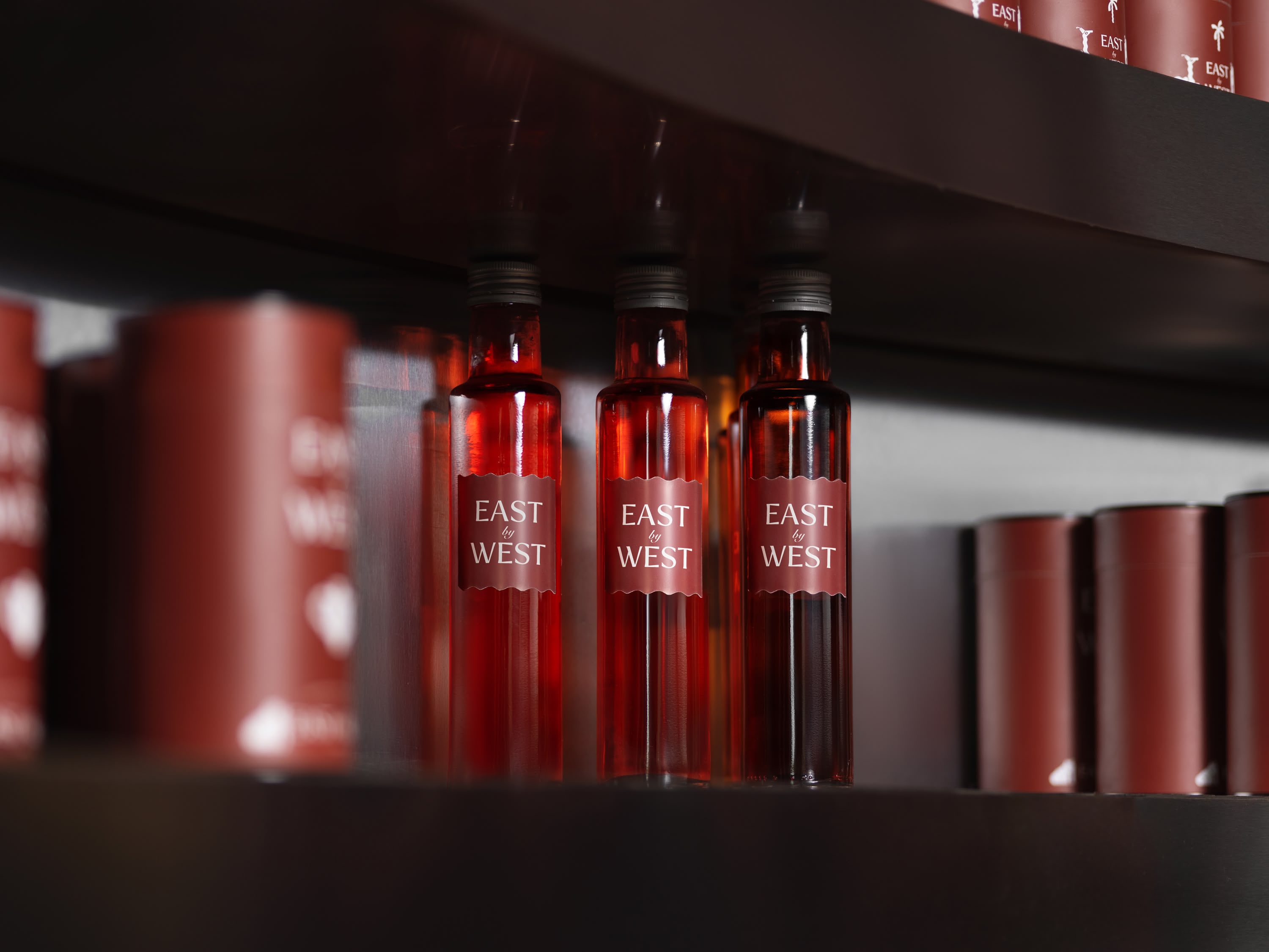 Three red glass bottles labeled "East by West" arranged on wooden shelf beside matching cylindrical containers in dimly lit display. 