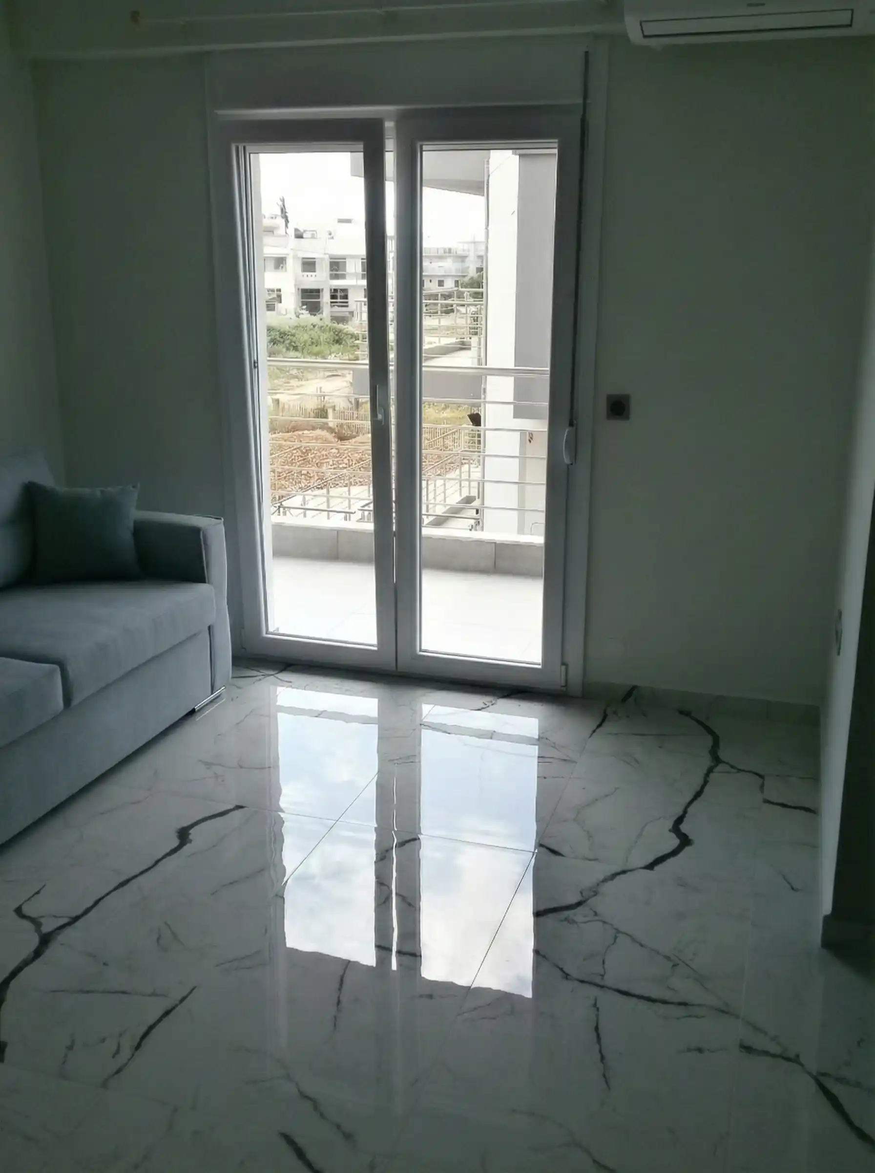 Μοντέρνο σαλόνι με γκρι καναπέ, μαρμάρινο δάπεδο και μεγάλη μπαλκονόπορτα που οδηγεί σε βεράντα.