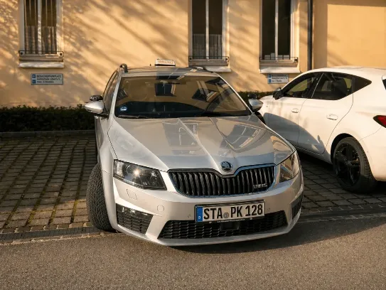Frontansicht eines silbernen Skoda Octavia RS Fahrschulautos mit dem Kennzeichen STA-PK 128 auf einem Parkplatz vor dem Fahrschulgebäude.