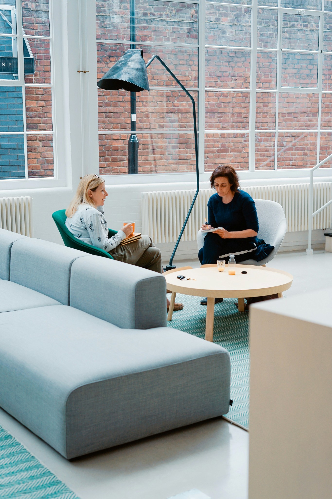 Two people are seated in a modern, well-lit office with large windows and exposed brick walls. One person is sitting in a green chair holding a cup and a notebook, while the other person is seated on a light grey chair, holding a clipboard and writing something down. Between them is a round wooden table with a water bottle, a glass of water, and some small objects on it.