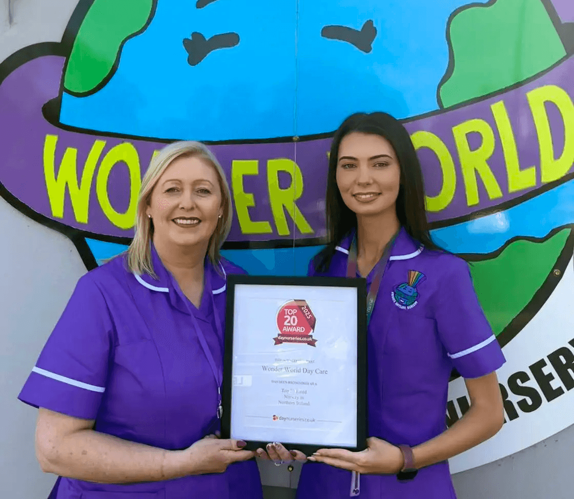 Two women in purple uniforms holding a framed award in front of a "Wonder World" sign.