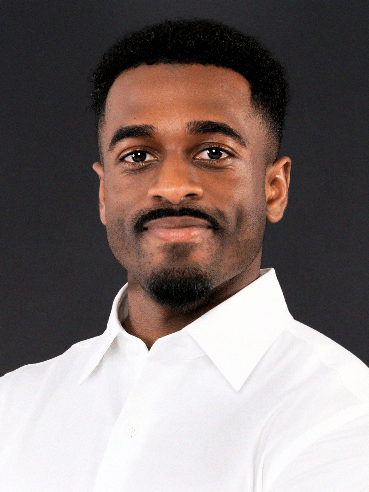Man with short hair and beard wearing a dark blazer and black shirt against a plain background.