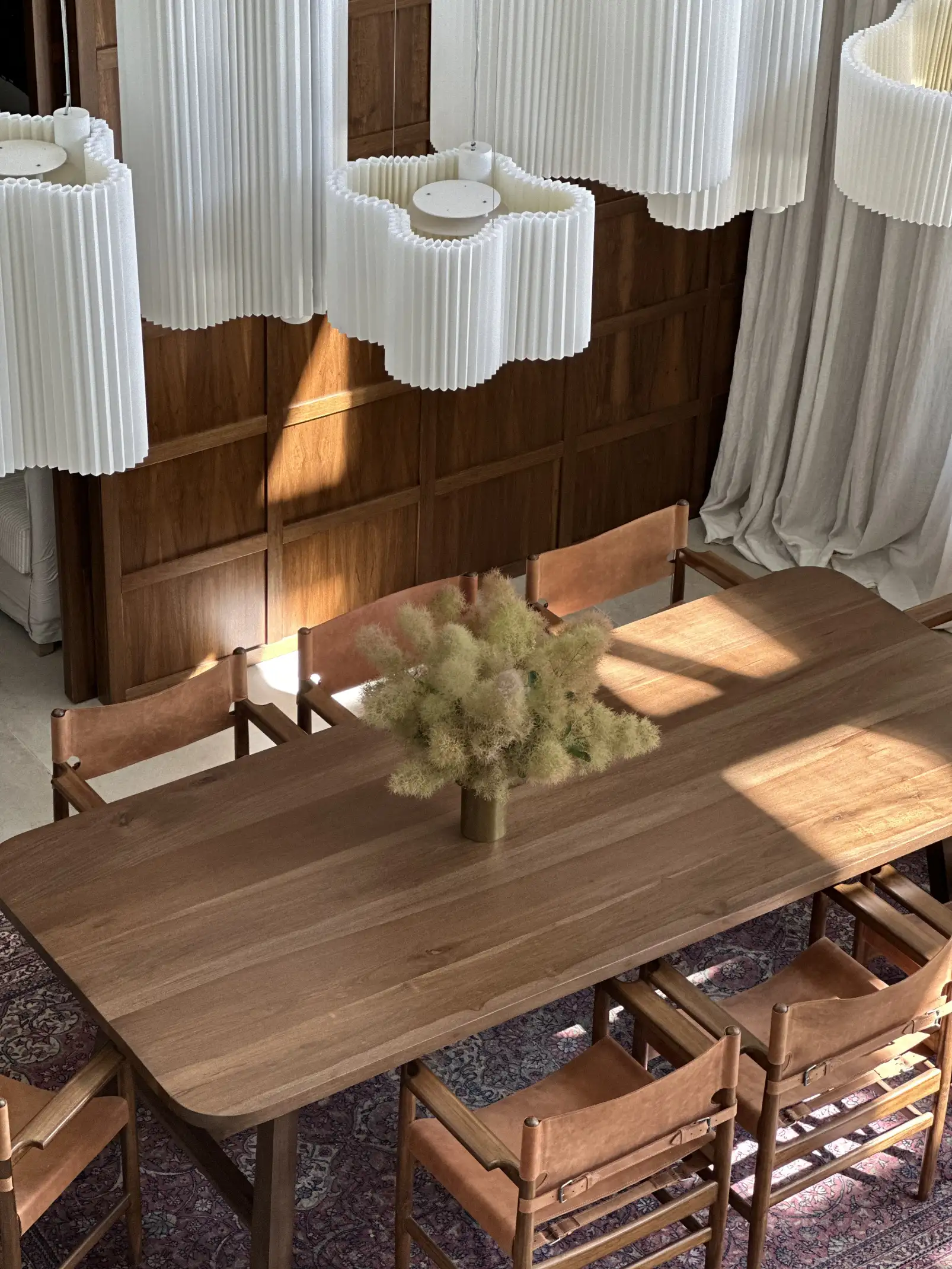 Vista superior de comedor de madera con lámparas plisadas y arreglo floral neutro para estudios de interiorismo que comunican armonía y textura natural.