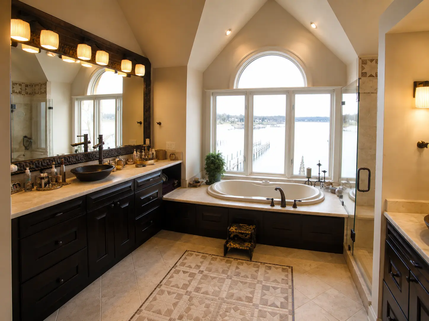 Luxury primary bathroom renovation with dark wood double vanity, soaking tub, arched window, and waterfront view.